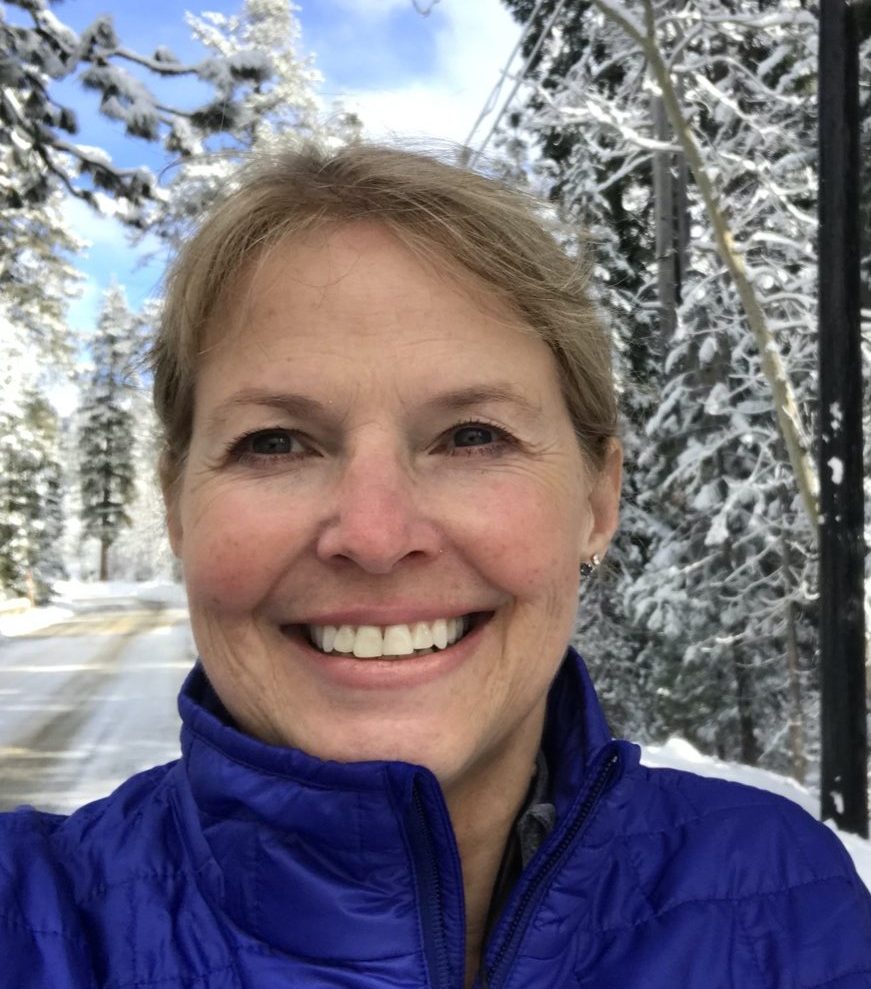 a woman with shirt blond hair smiles at a camera. she's standing in a beautiful snowy landscape