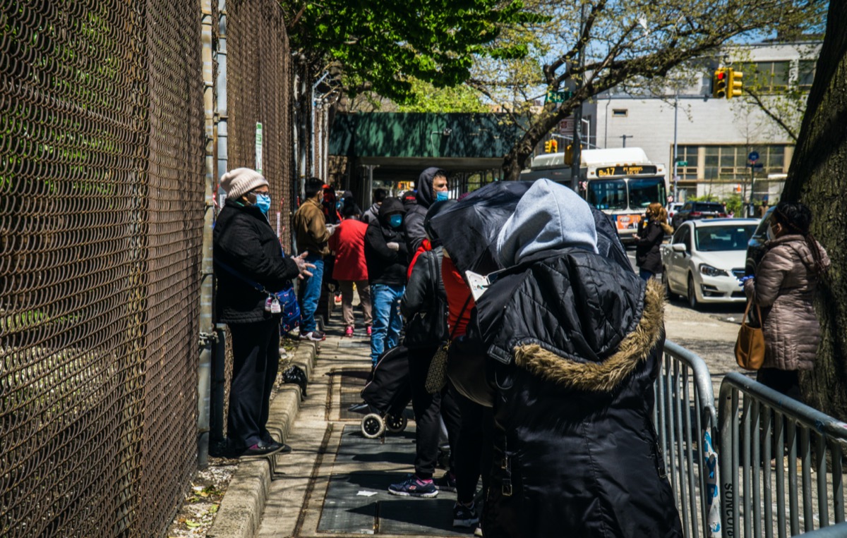 people of color wearing face masks line up outside of a building along a wire fence