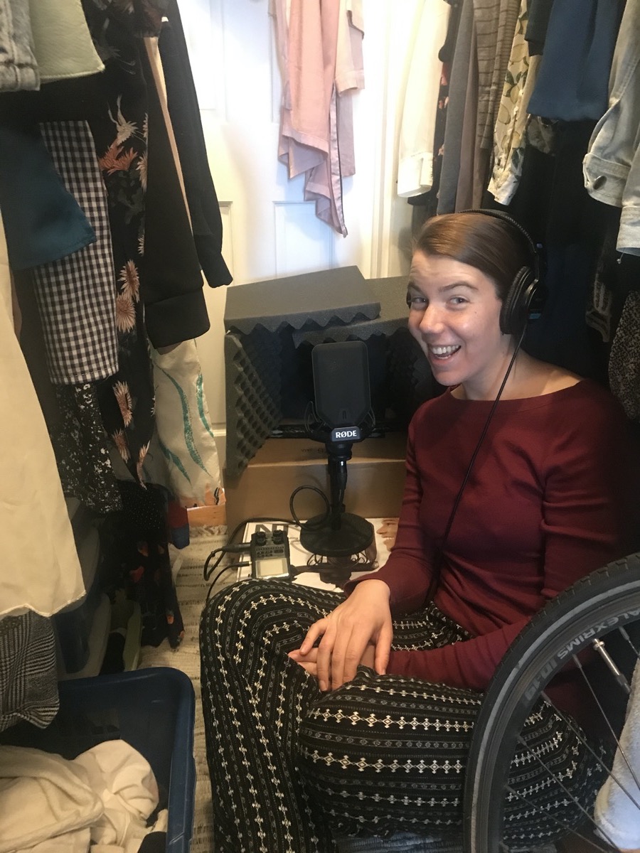 a woman sits in a closet with headphones speaking into a microphone propped on a box surrounded by foam blocks