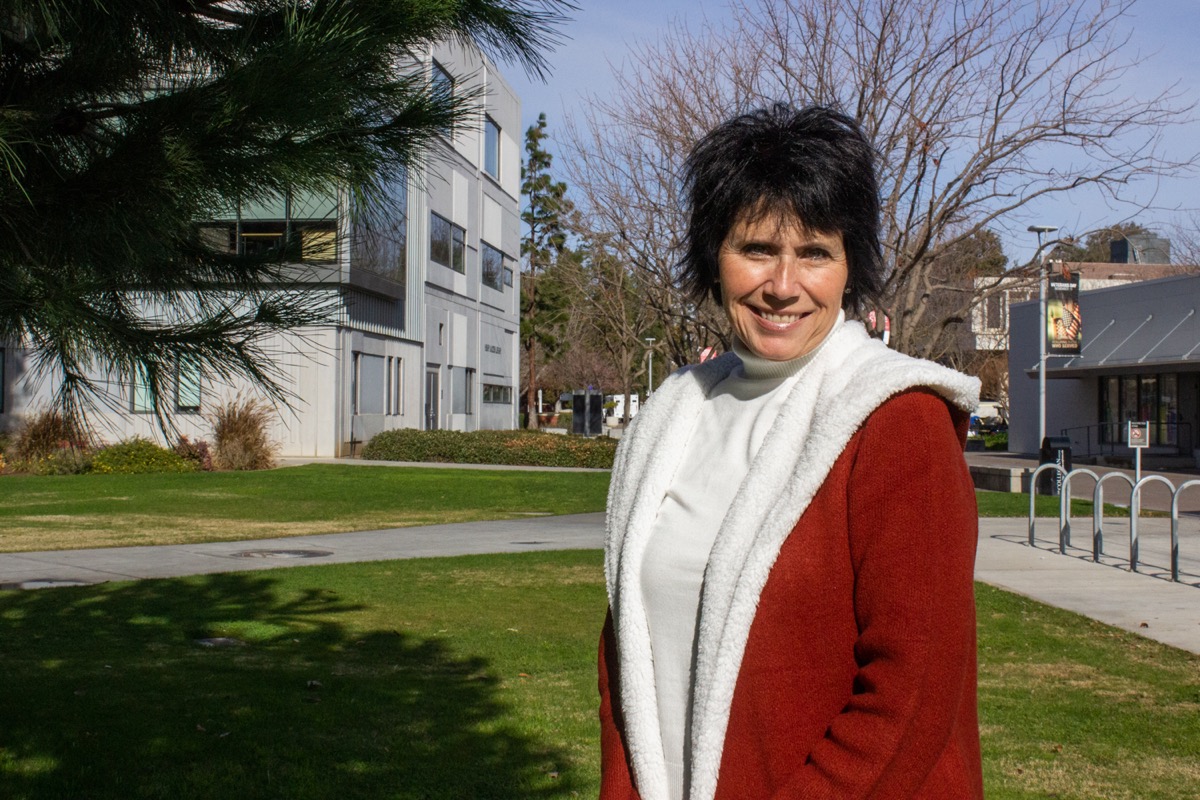 a woman with short black hair and a red coat stands outside on a grassy lawn smiling at the camera