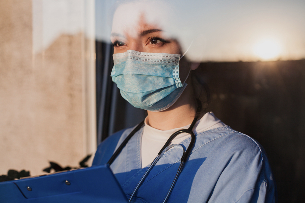 a female medical care worker in scrubs and a face mask, holding a clipboard. she looks out a window with a furrow brow, as the sun gleam reflects off the surface
