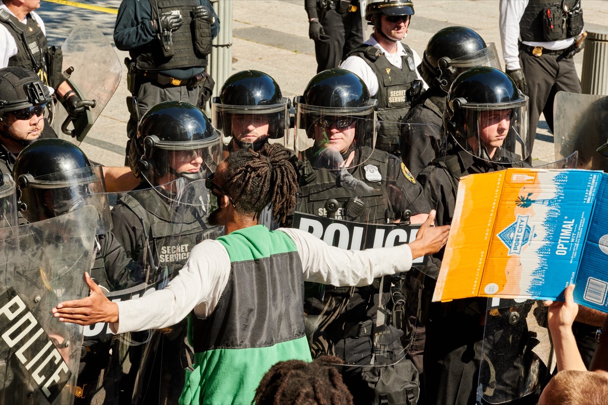 a black man peacefully protesting with his arms spread out wide. he stands in front of a wall of police with shields. a large crowd stands behind him