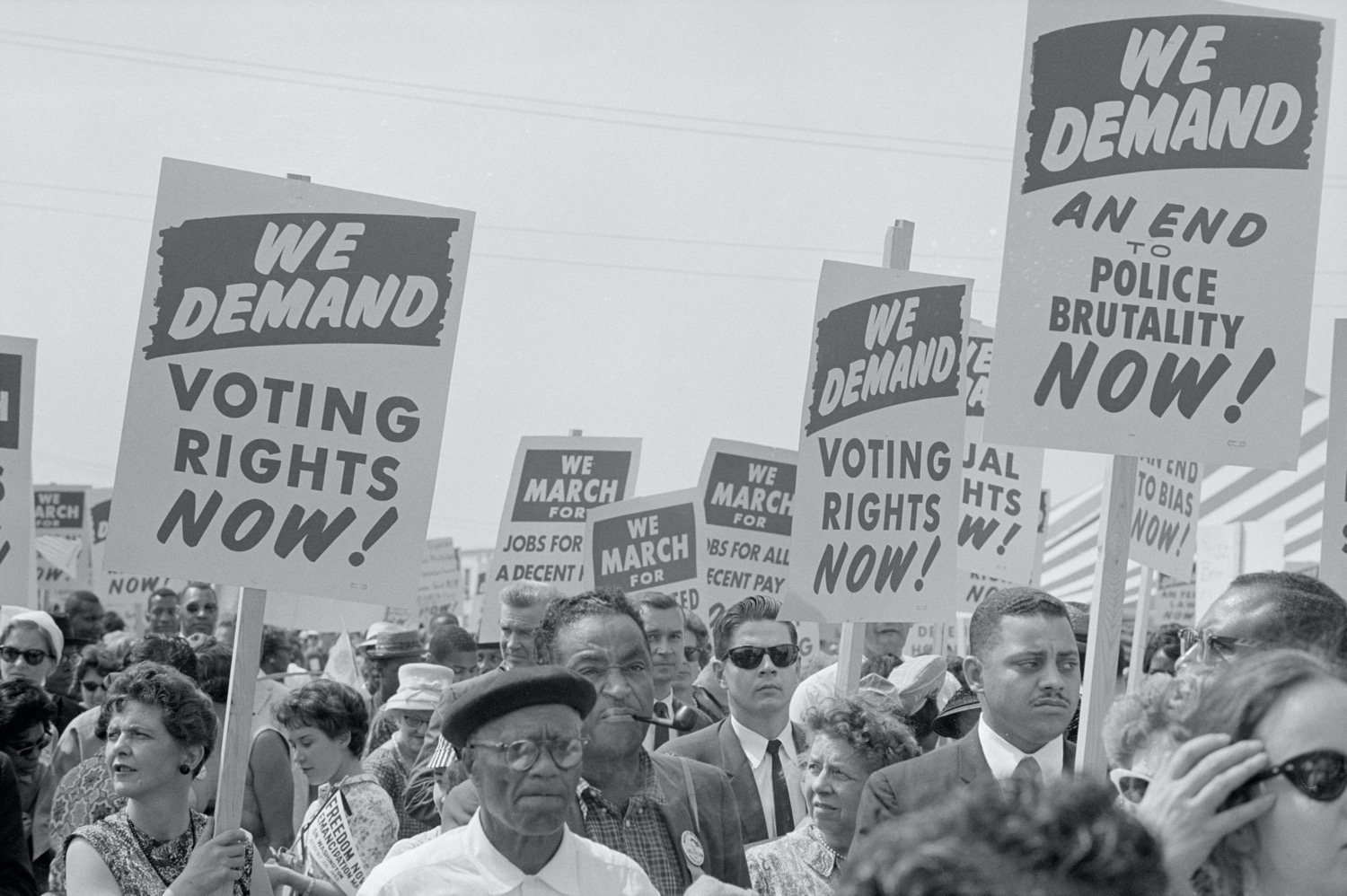 black and white photo of people marching with signs that say "we demand voting right now, we demand an end to police brutality, we demand voting right now"