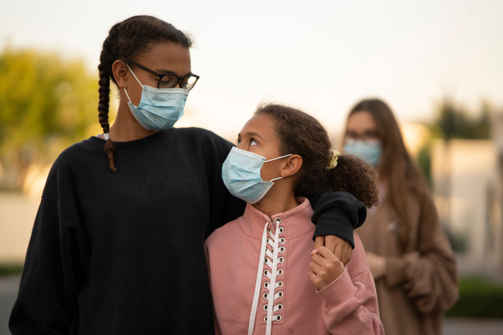 two Black teenaged teens wearing face masks looking at each other fondly while walking embraced