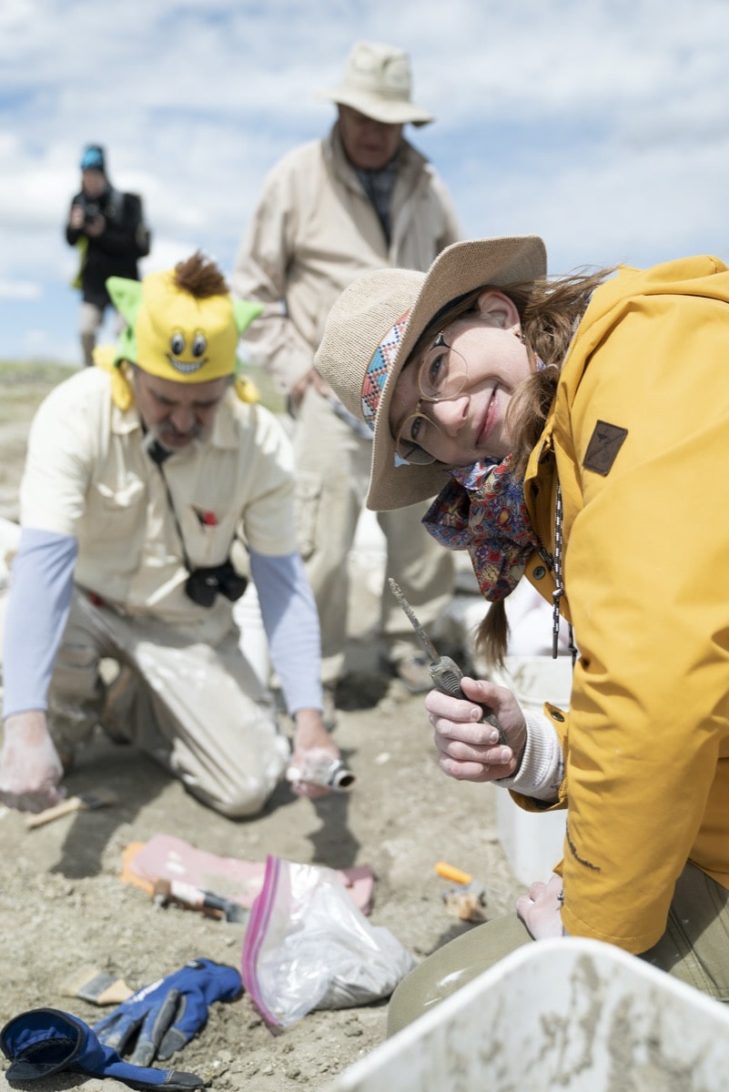 in focus a woman in a straw hat and yellow jacket holds up a digging tool, in the background are paleontologists digging at a fossil site