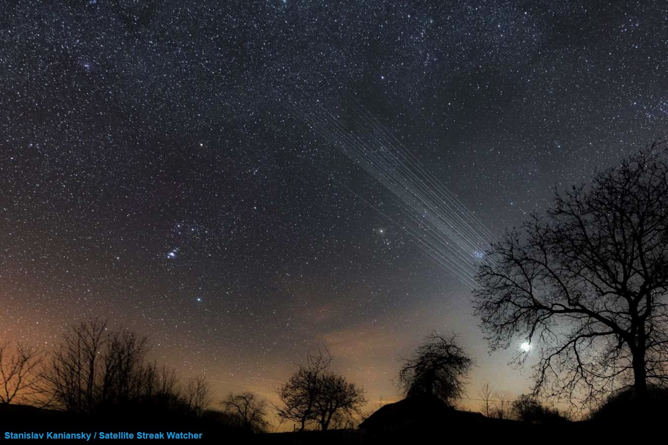 the sun has set giving off a faint orange glow and an open night sky above a treeline. you can see stars, but white streaks can be seen in the left corner of the image