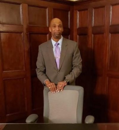 a black man in a suit stands behind a desk chair in a wood paneled office