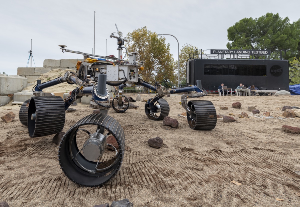 a rover robot with six large wheels out on a sandy field. you can see the wheel tracks in the sand