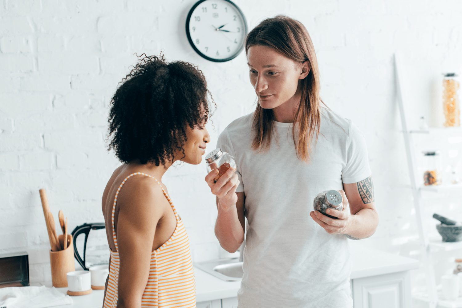 young man with tattooed hand giving girlfriend a sniff of spices at kitchen