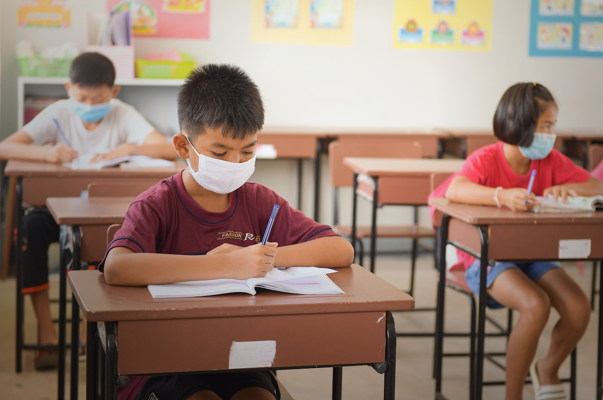 a young asian boy with a face mask writes in a notebook in a classroom. two other students are in the class seated several desks away