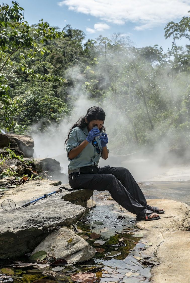 a woman scientist wearing gloves looks a specimen through a microscope as she sits on rocks along a steaming river