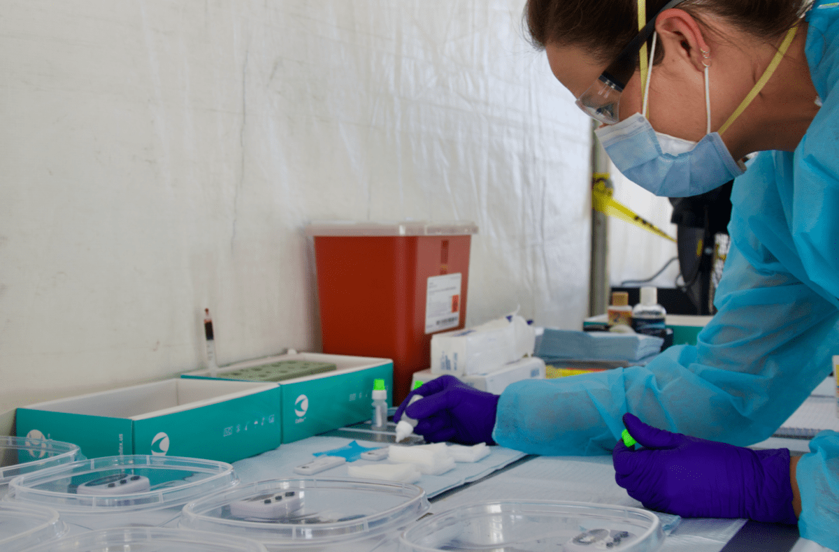 a nurse in a makeshift tent wearing protective gear readies multiple test kits in clear plastic containers