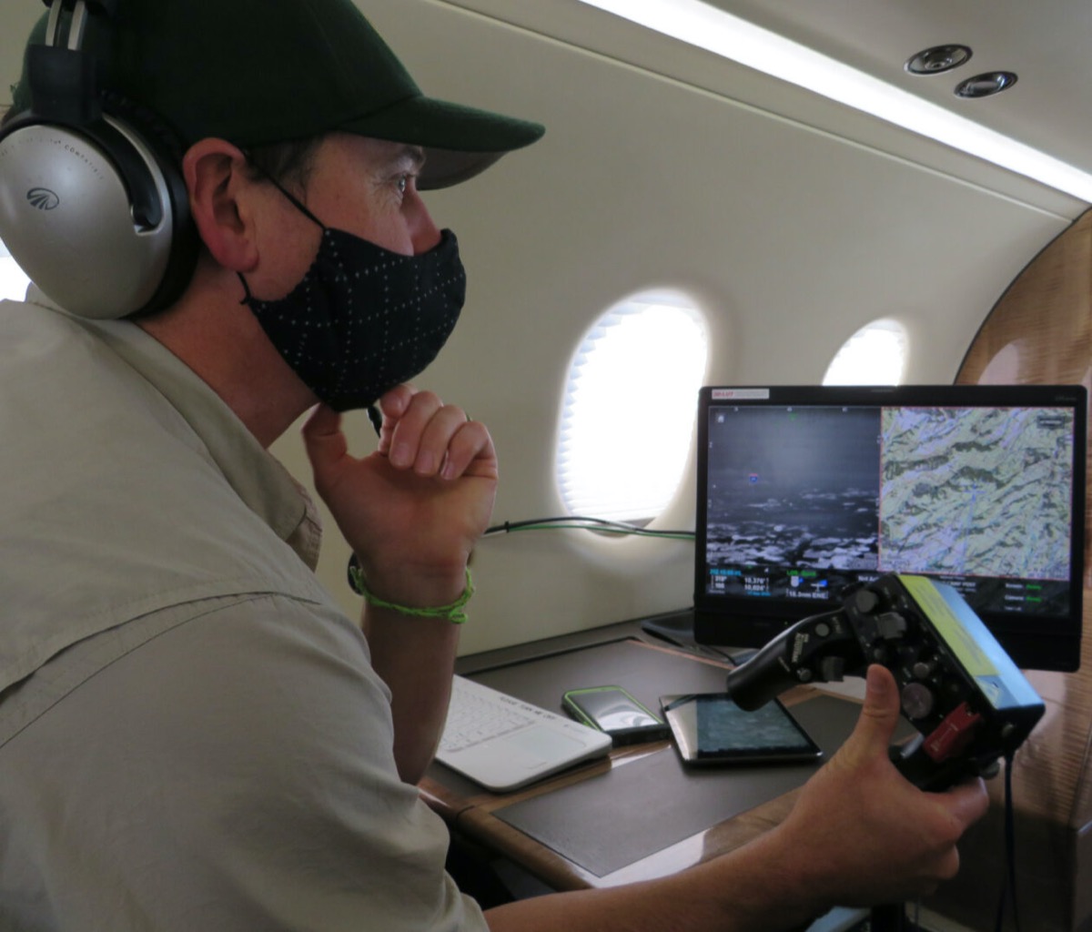 a man wearing a face mask holds a video game-like remote control and looks at a computer screen of maps. he's inside of a plane cabin