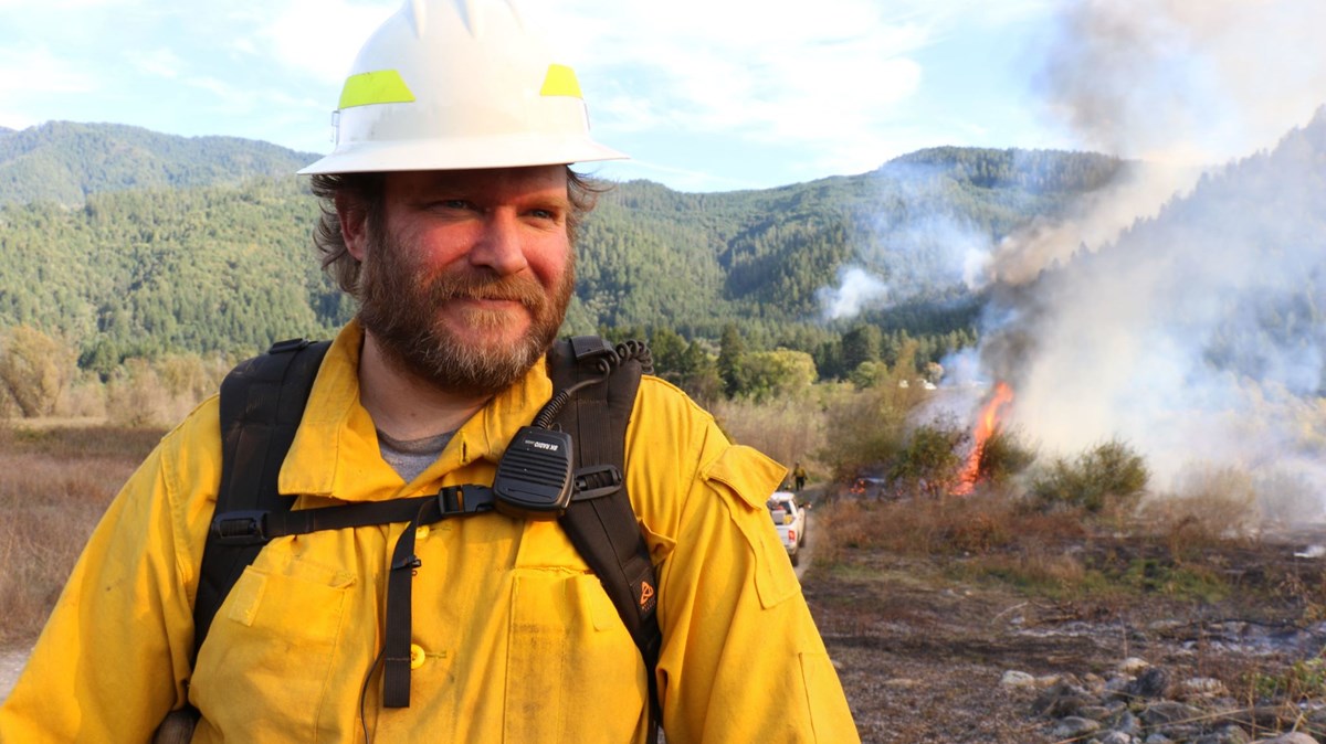 an indigenous man in a yellow safety jacket and hard hat with small fires burning behind him