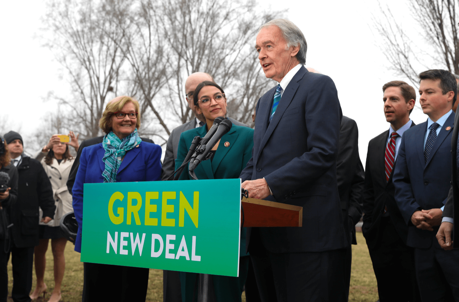 ed markey stands at a podium that says "green new deal" flanked by people to his right, including alexadria ocasio-cortez