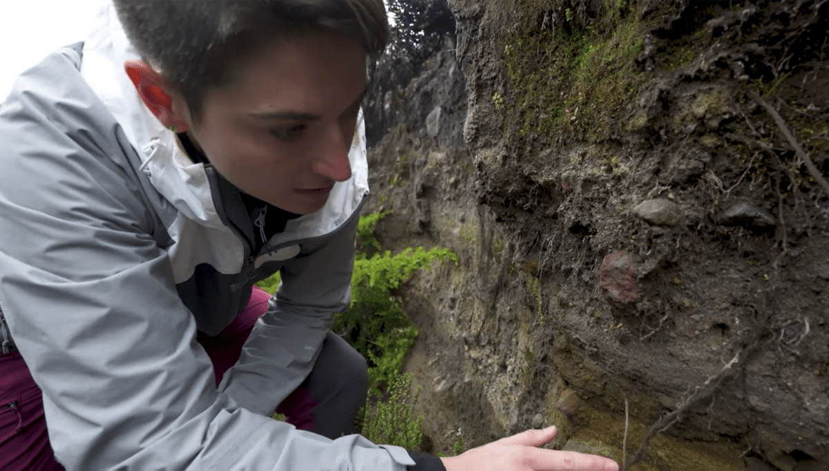 a woman crouches next to a wall of rock and runs her hand along the layers