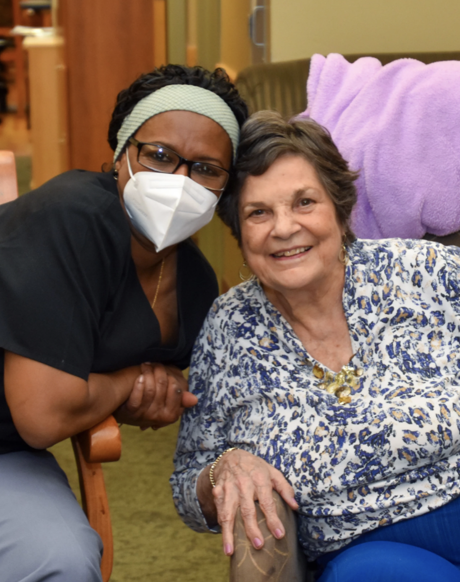 a nurse in a mask crouches next to an older lady in a nursing home