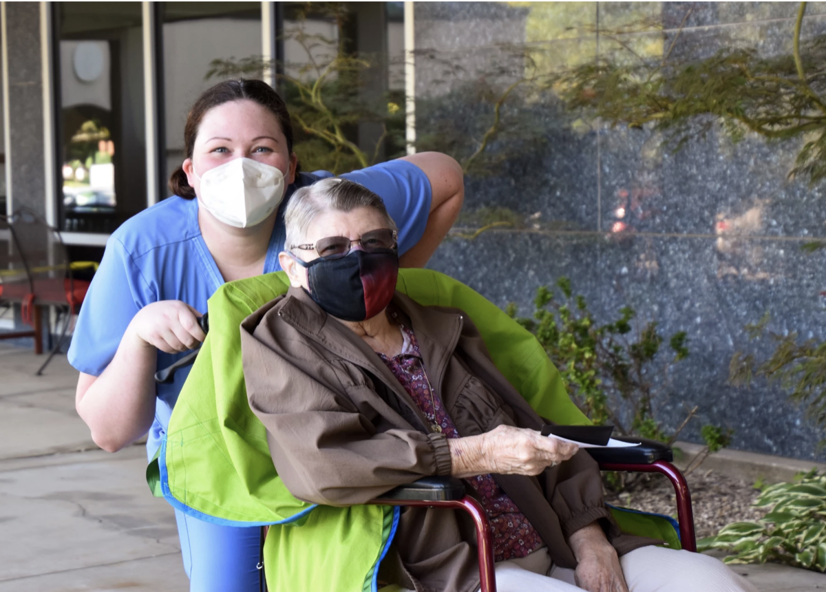 a nursing aid with a mask on pushing an older woman in a mask in a wheelchair.