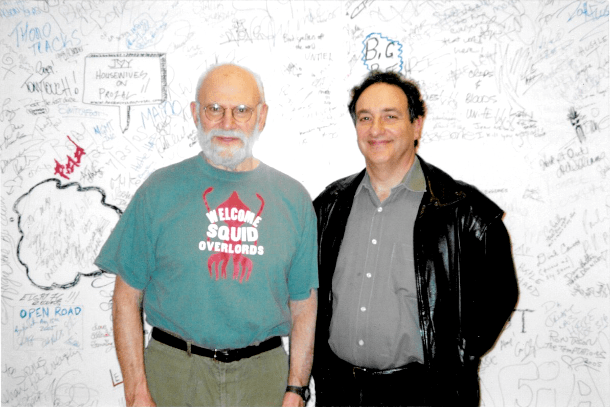 oliver sacks and ira flatow smiling at the camera in front of a wall filled with handwriting