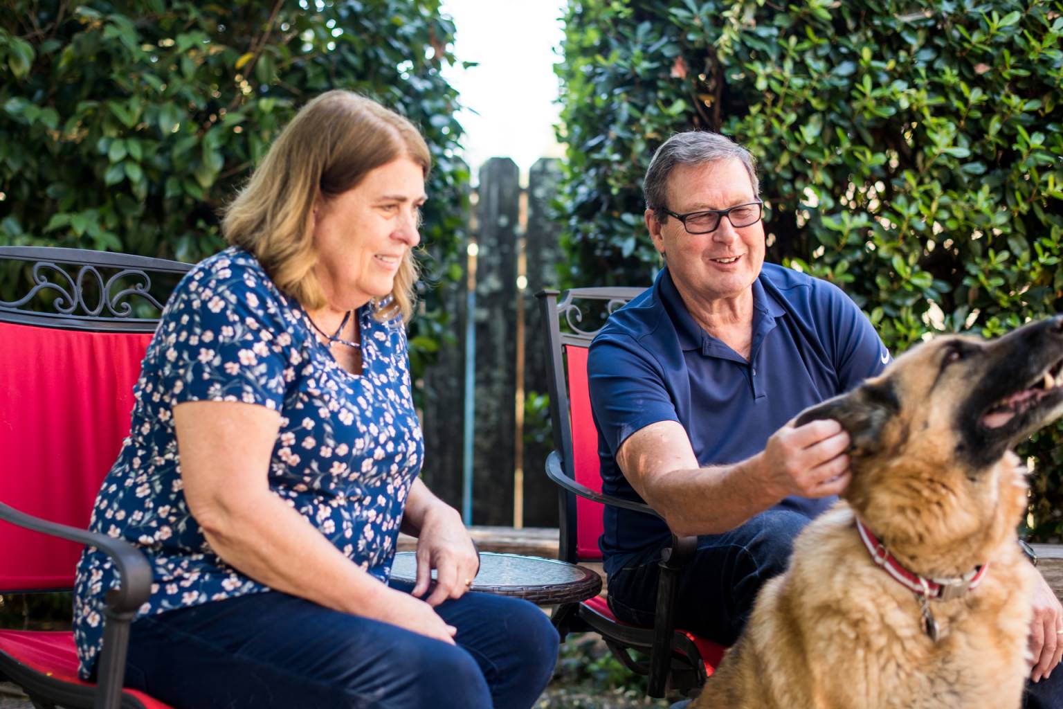 a woman and a man sitting in red chairs outside petting a dog