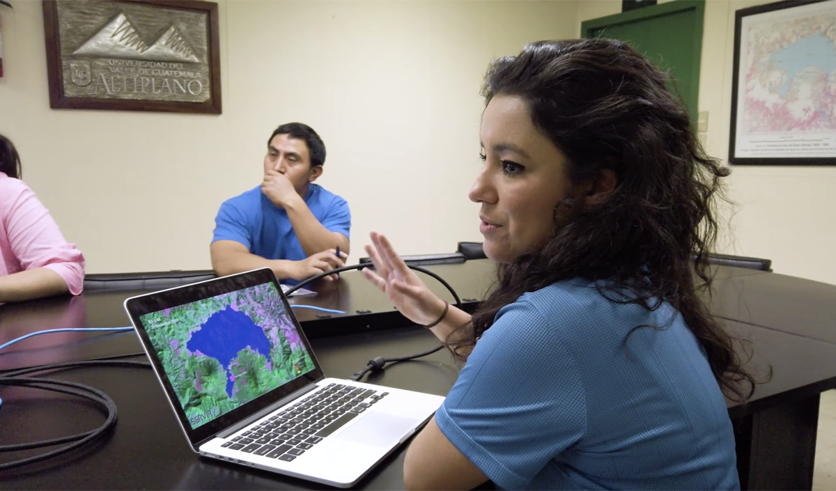 a woman sits in front of a laptop with a satellite image of a lake and she speaks to a table of fellow scientists