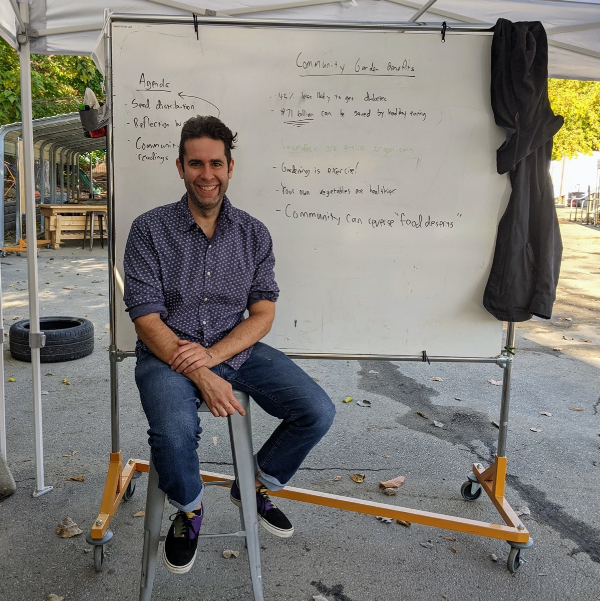 a man sitting on a stool outside in front of a white board. it's an outdoor classroom!