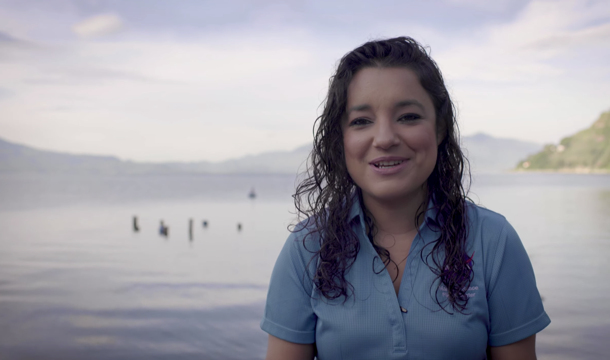 a woman with wavy hair and wearing a blue polo shirt sits in front of a beautiful lake. she's smiling