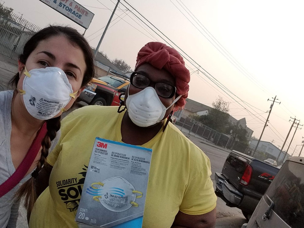 a Latino woman and a Black woman wearing face masks pose for the camera with smoke in the background