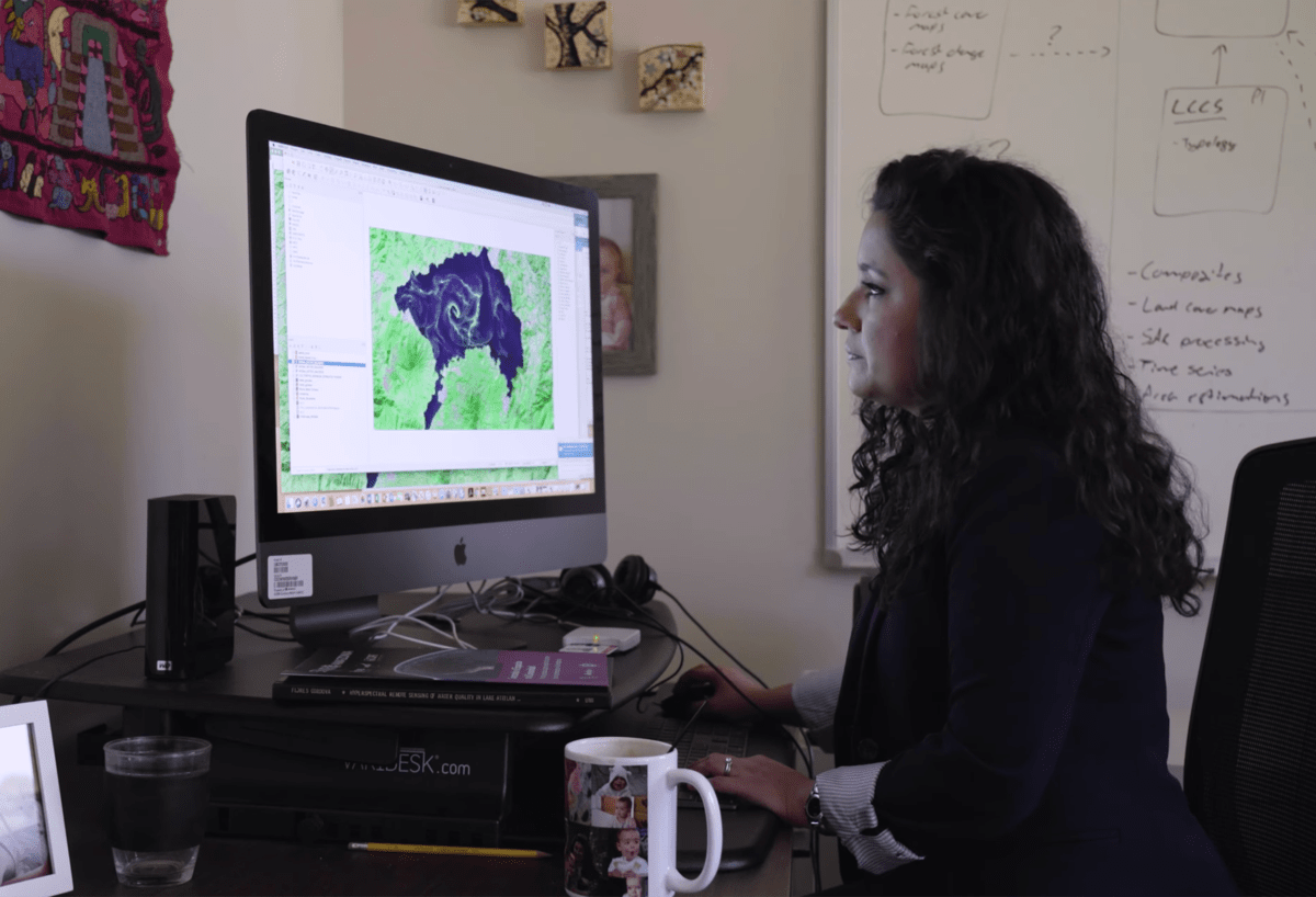 woman in a blazer in her office looking over a computer monitor with a satellite image of a lake experiencing an algal bloom