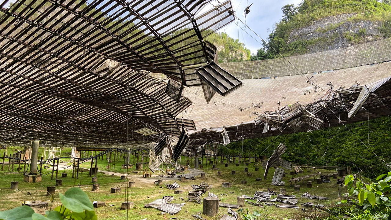 the underside of a massive dish structure, where metal panels have fallen off and some are hanging from the dish