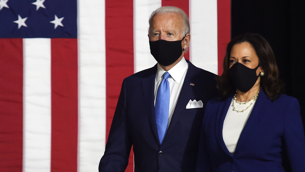 biden and harris, wearing masks, walking on stage, with an american flag in the background