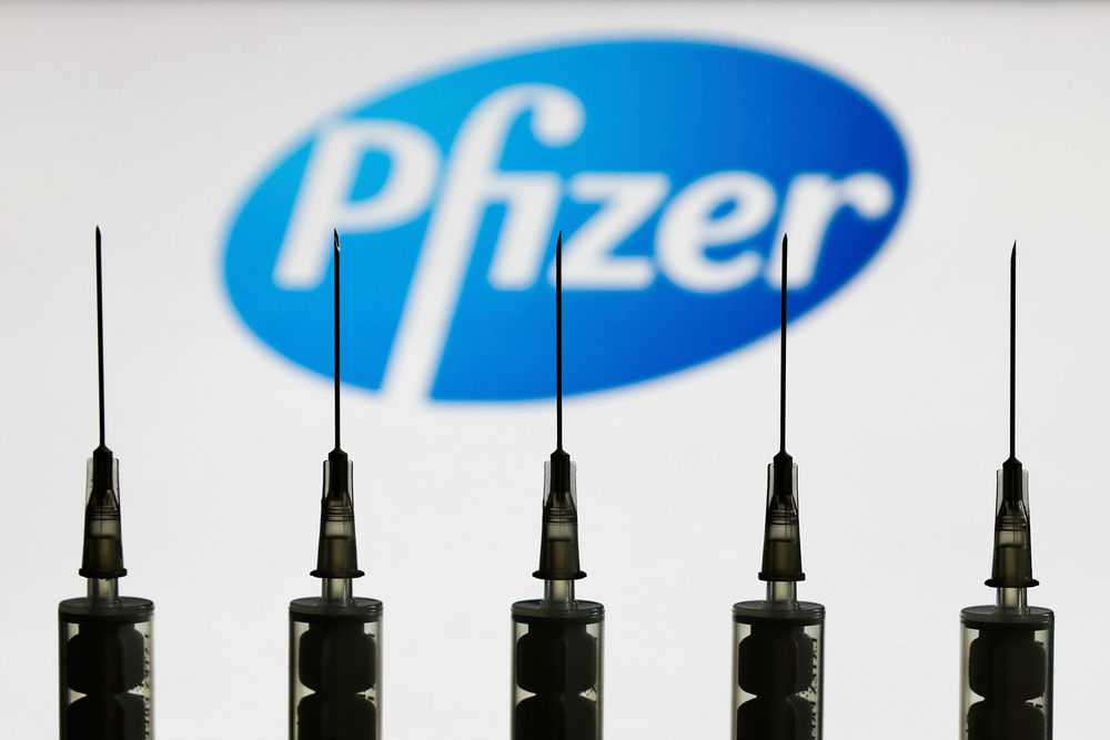 a close up of five needle points of syringes lined up in front of a blue logo of the word pfizer in the background
