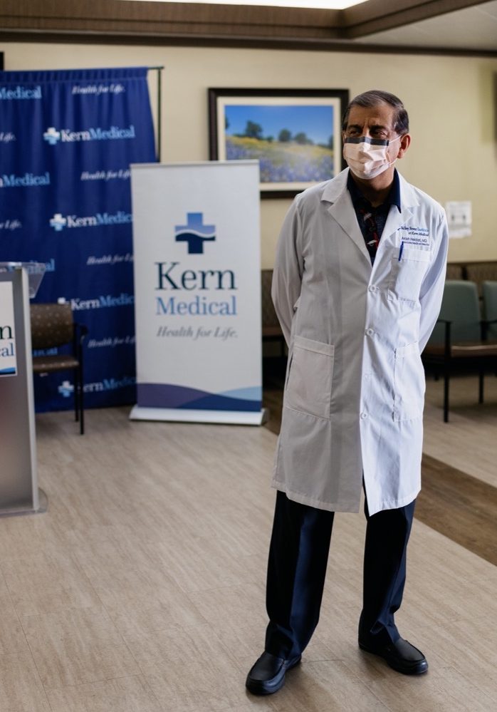 a male doctor wearing a white lab coat and face mask stands in a conference hall. behind him is a poster that reads "kern medical"