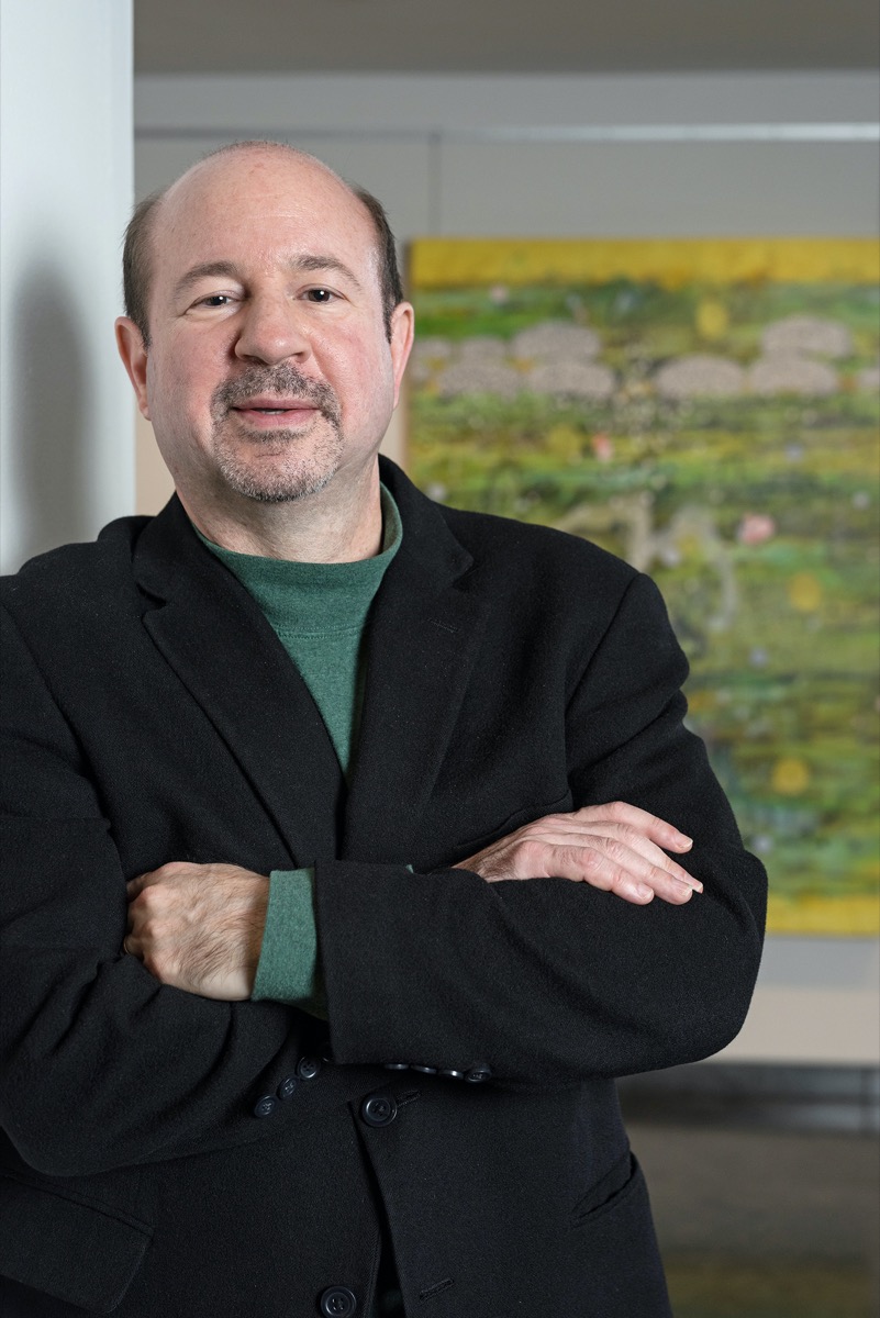 a white man leaning against a wall in front of a painting with his arms crossed over his chest