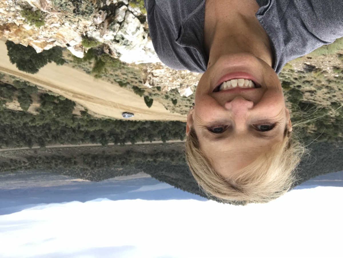 a woman taking a selfie on a hiking trail. behind her is a tree dotted valley and mountains