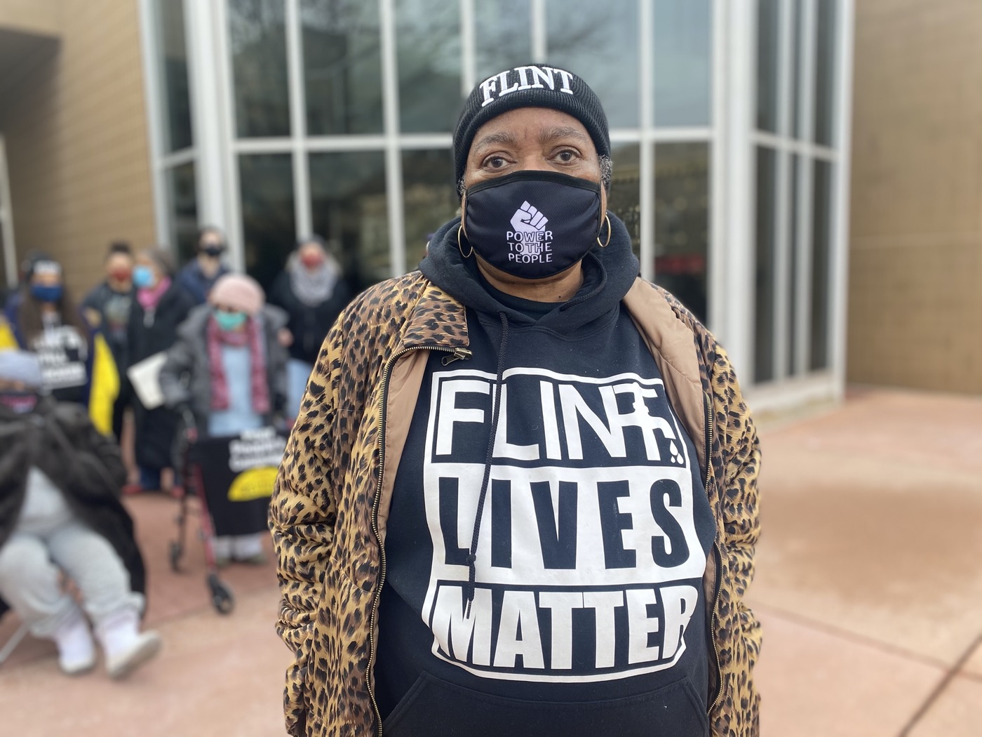 a Black woman protester wearing a beanie that says "flint", a mask that says "power to the people", and a shirt that says "flint lives matter"