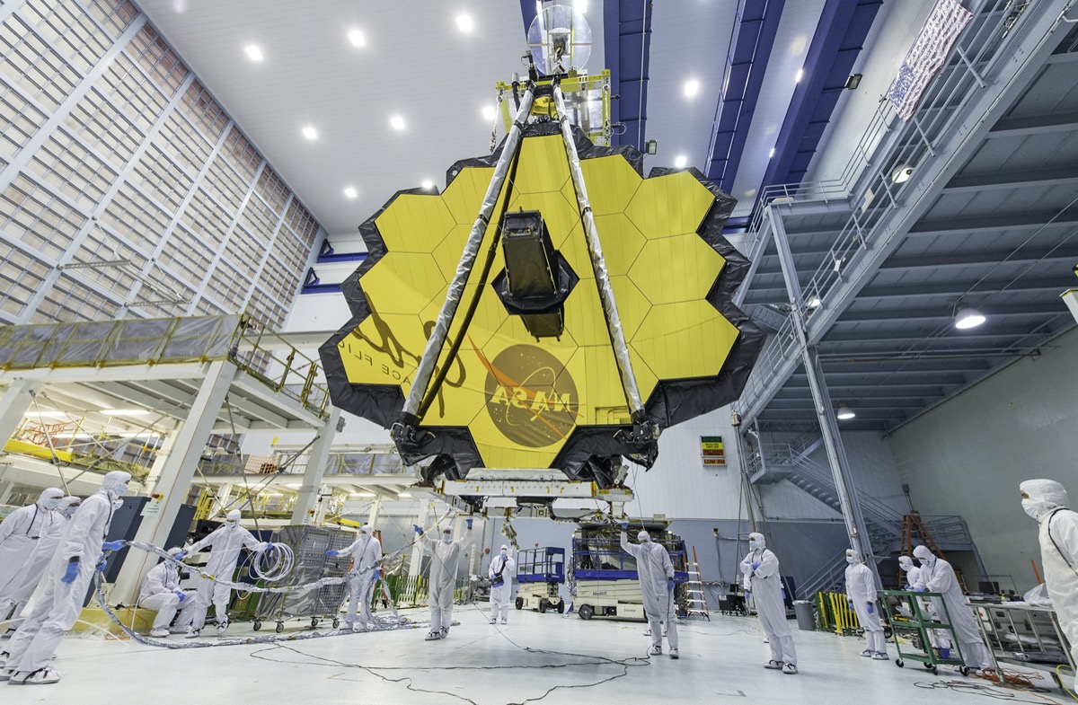 a giant gold disc made up of dozens of gold octagons in a massive hanger clean room with scientists in white protective suits around it