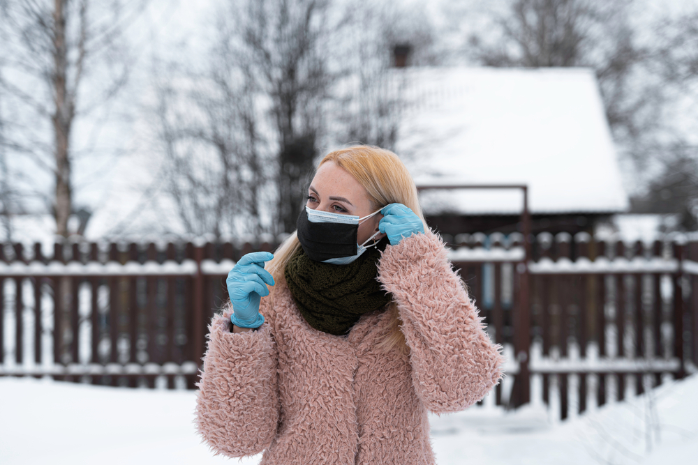 a White woman outside in a coat wearing gloves applying two masks to her face, and there's snow on the ground