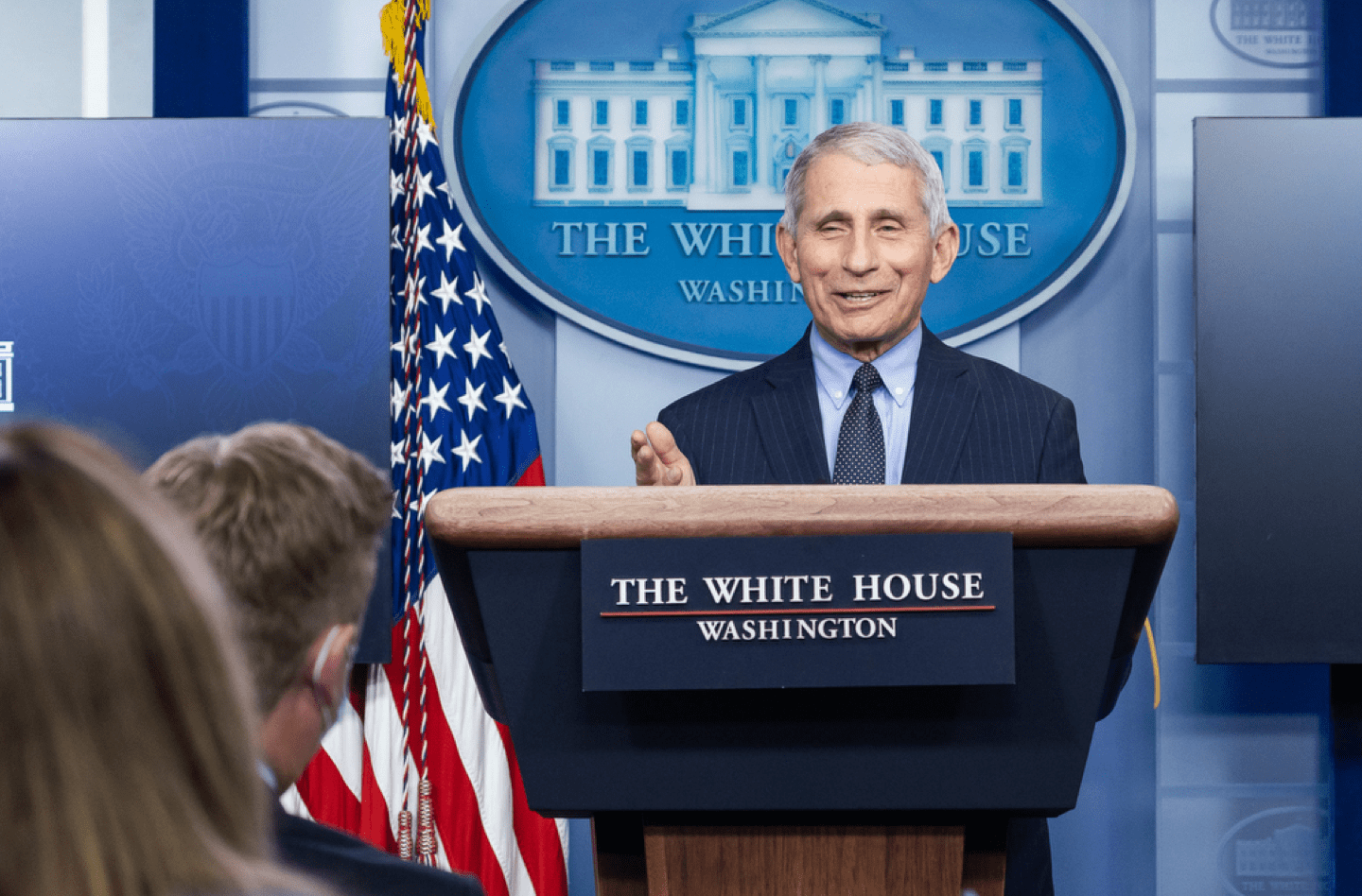 a smiling fauci speaks behind a podium in the white house briefing room