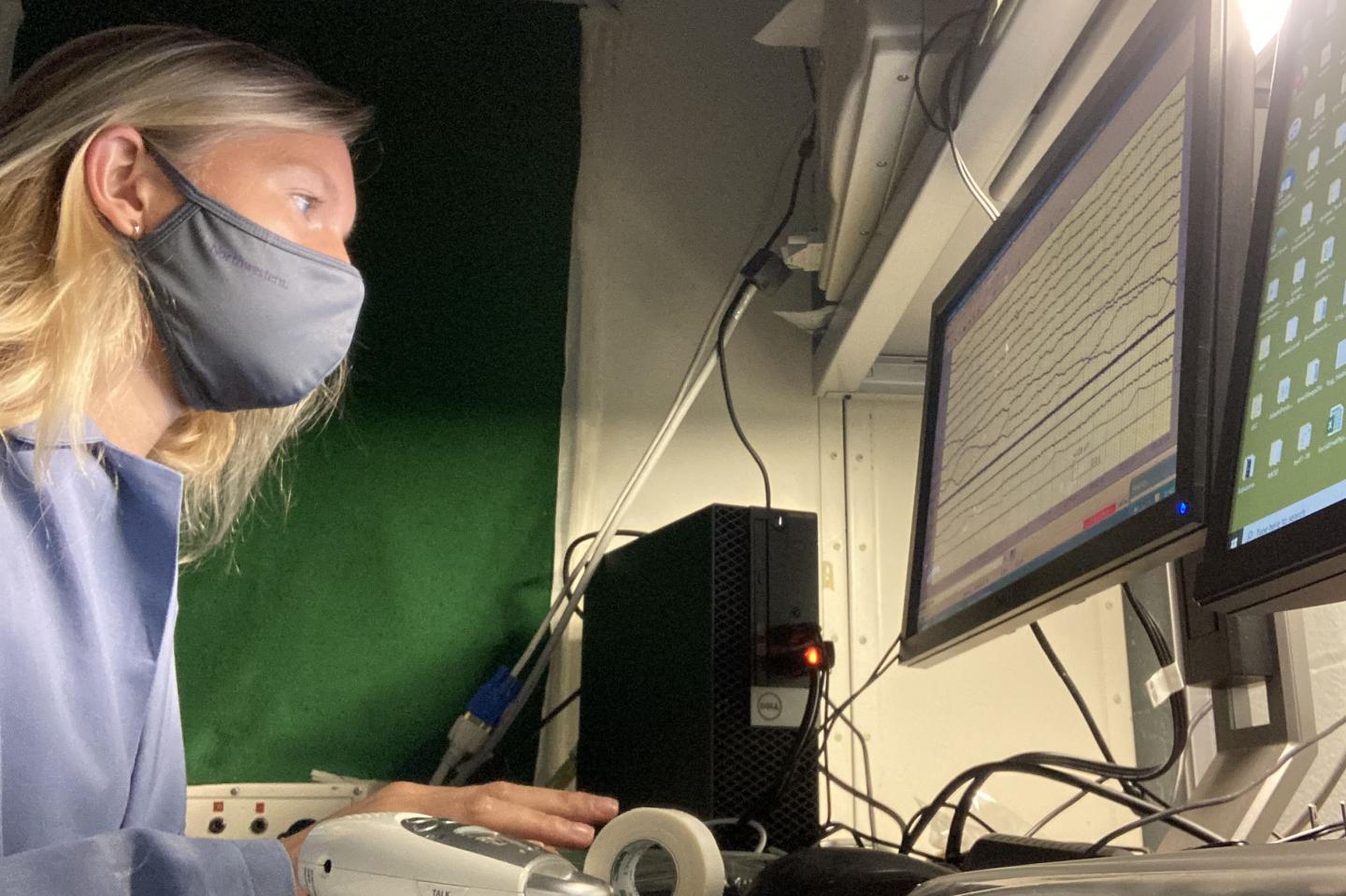 a woman in a face mask at a computer monitoring a graph of lines running horizontally across the screen