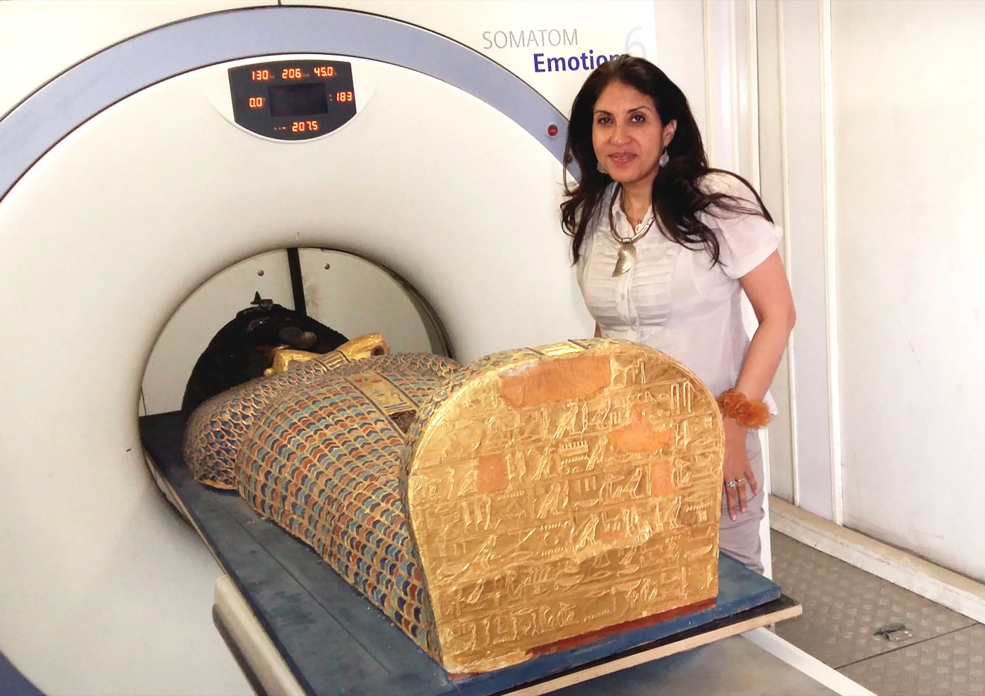 an Egyptian woman stands next to a CT scanner, which has an ornately decorated sarcophagus on its bed