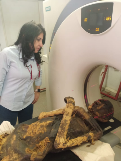 an Egyptian woman stands over an unwrapped mummy on a ct scanner. the skin is brown and dark brown, and the mummy's arms are crossed over its chest in an x-position