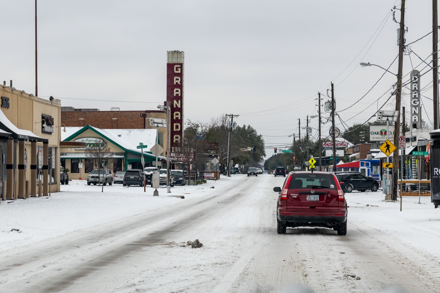 a snow covered street in dallas texas. lots of ice still on the street as a car drives down the street, lined with shops and a movie theater. no electric lights are present