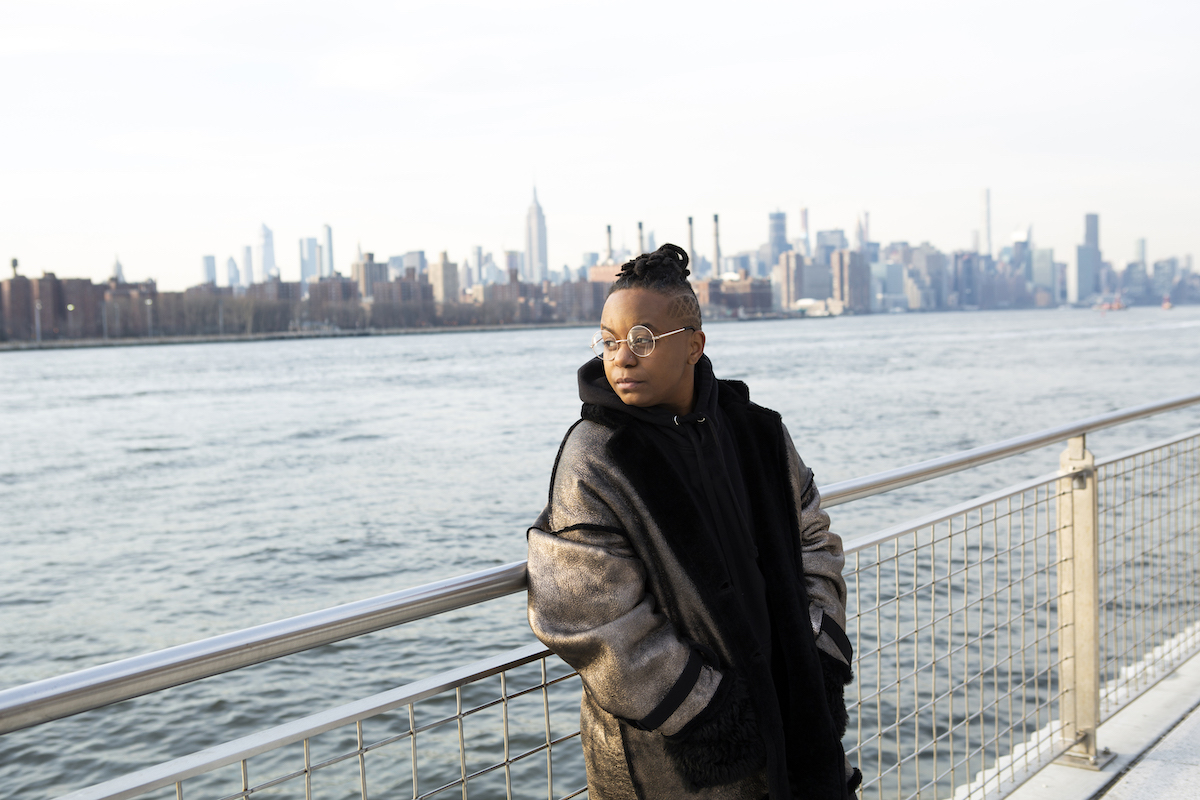 a black transmasculine person standing alone by the water's edge with the nyc skyline in the background, looking out into the distance