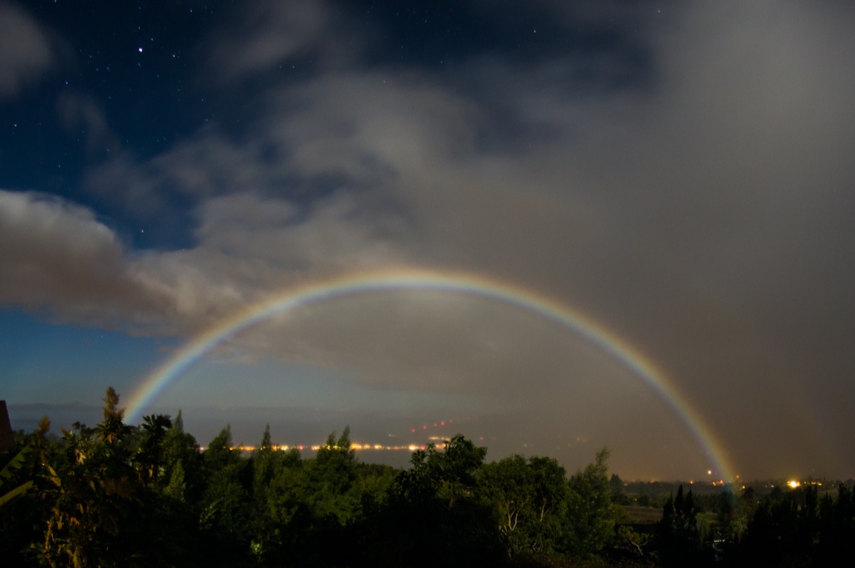 a faint rainbow seen arching over trees and city lights at night. you can see stars in the background