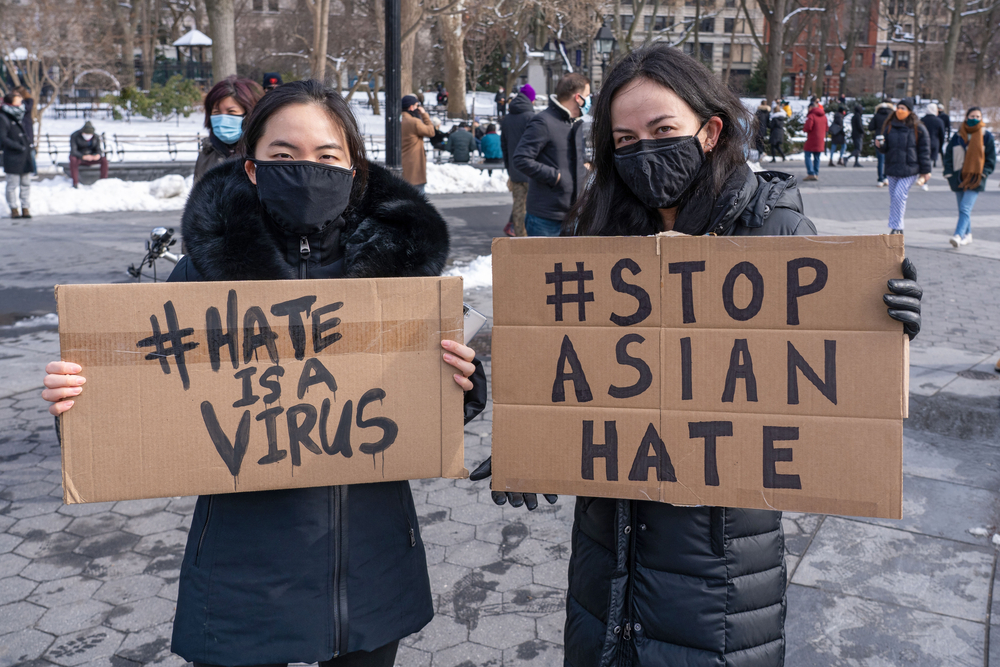 two asian women wearing masks and jackets outside in the snow carrying cardboard signs that say 'hate is a virus' and 'stop asian hate'