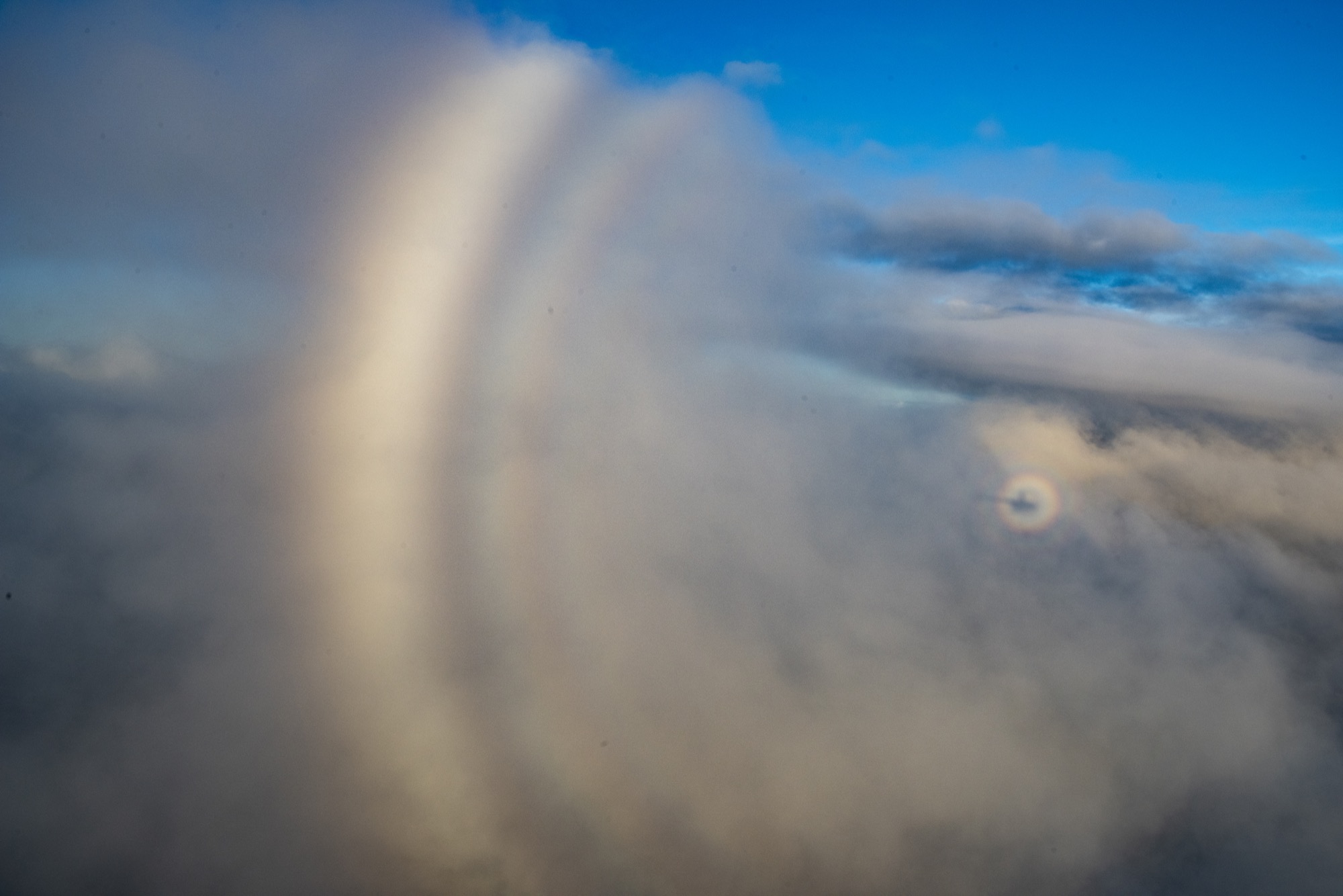 a photo taken from a helicopter. in the distance you can see the shadow of the helicopter against the clouds, around the shadow is a glowing circular rainbow ring. to the left is another partial curve that is a cloud bow
