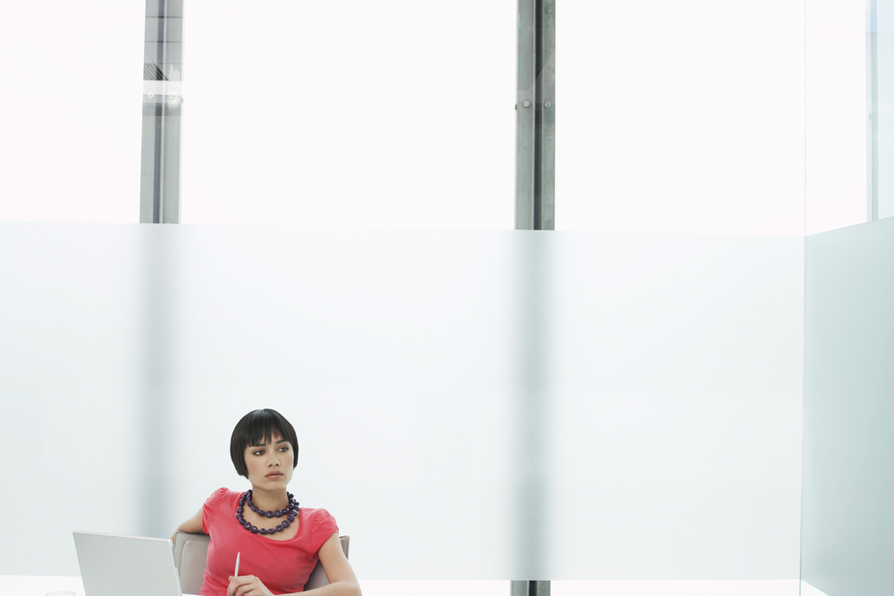 a woman thinking and staring off in the distance as she sits with a laptop in a modern cubicle office