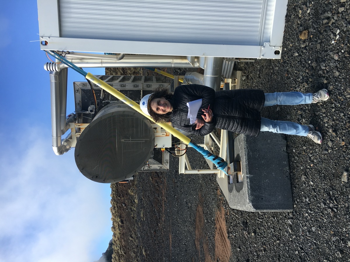 a woman wearing a hard hat and a winter coat holds a packet of paper and stands in front of industrial-looking equipment out on dark gravel, rocky terrain