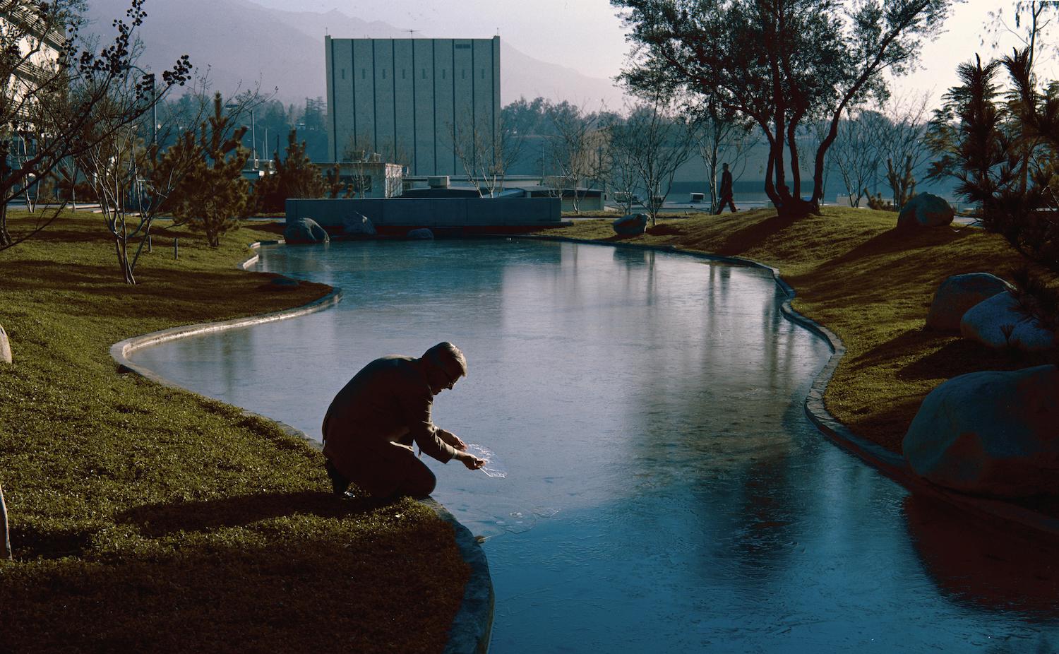 a winding human-made river cutting through grassy hills. in the background is a square white building and in the foreground, shadowed, is a person crouching down near the water